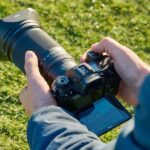 Суперзум Nikkor X 28–400 мм, прикрепленный к камере Nikon, сфотографирован через плечо фотографа на спортивной площадке.